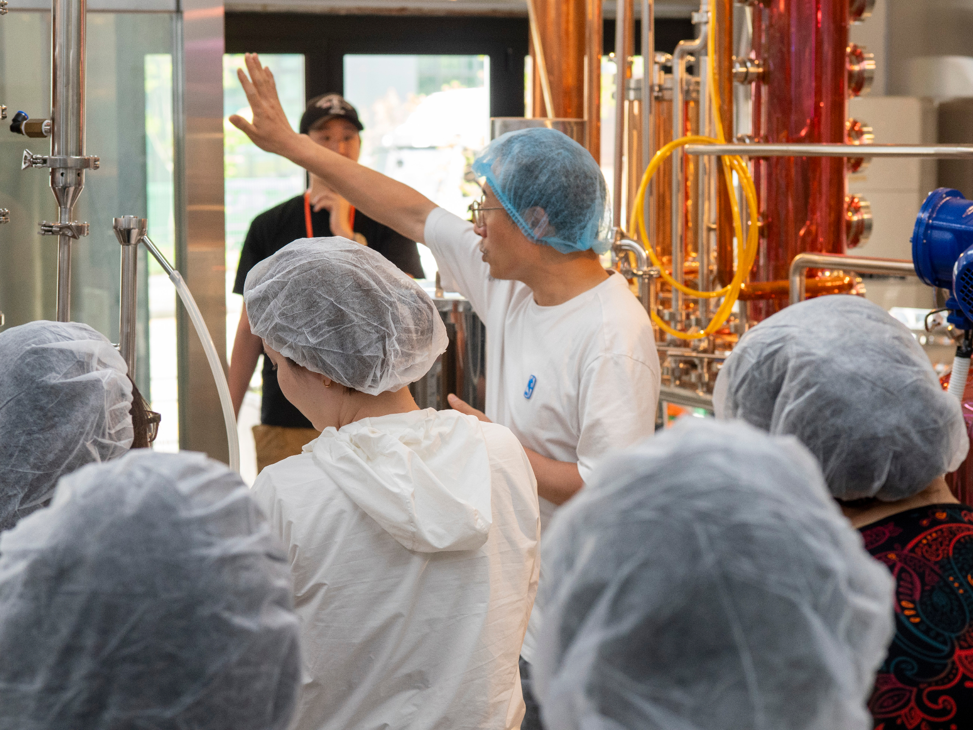 Modern distillery tour with visitors in protective gear observing copper distillation equipment